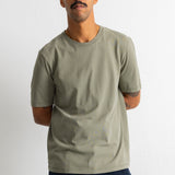 A man wearing a plain crew-neck t-shirt in a muted sage green color, paired with dark blue pants, standing against a light gray wall.