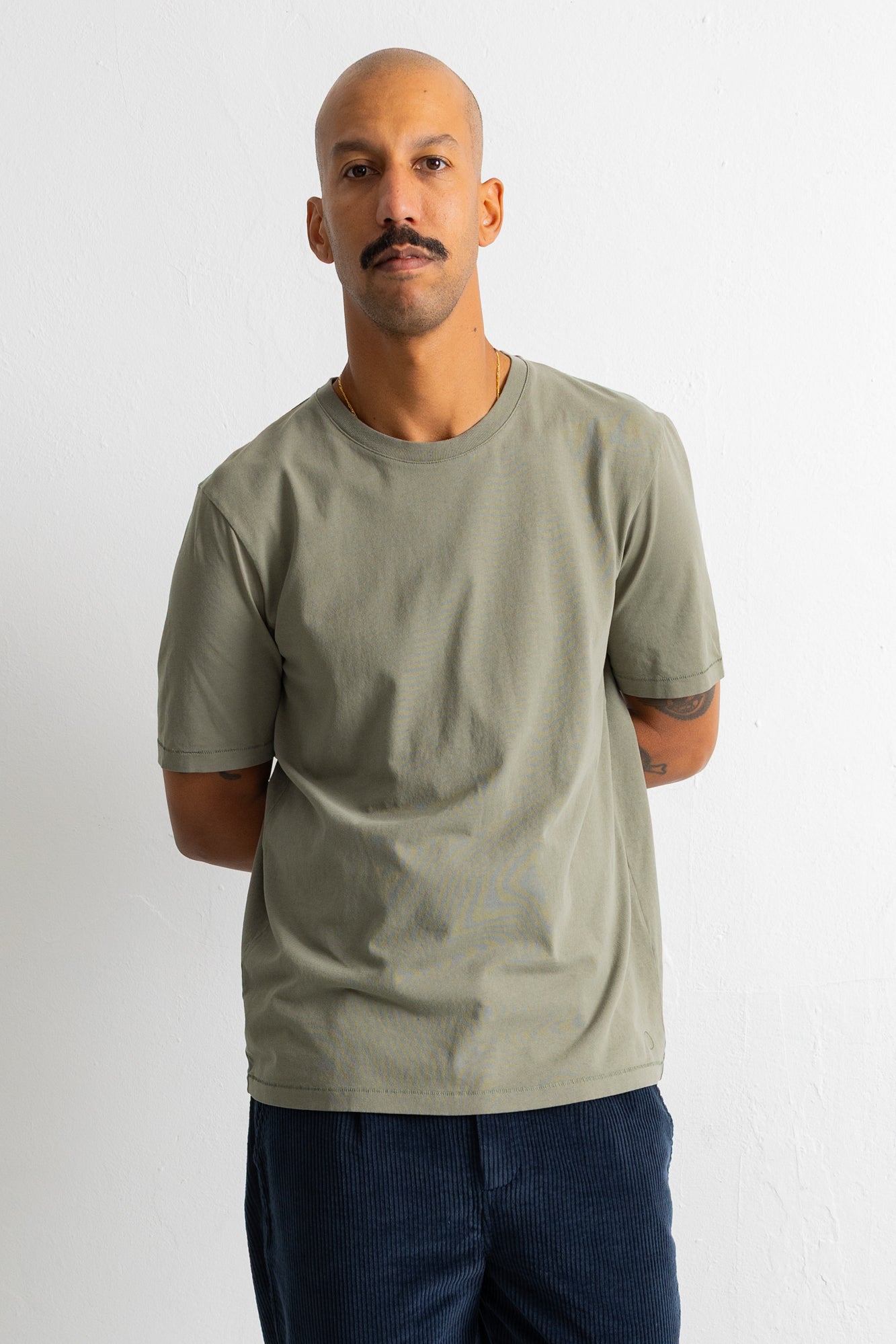 A man wearing a plain crew-neck t-shirt in a muted sage green color, paired with dark blue pants, standing against a light gray wall.