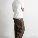 A man shown from the back wearing a white t-shirt, loose brown pants, and dark sneakers, standing against a plain white wall.