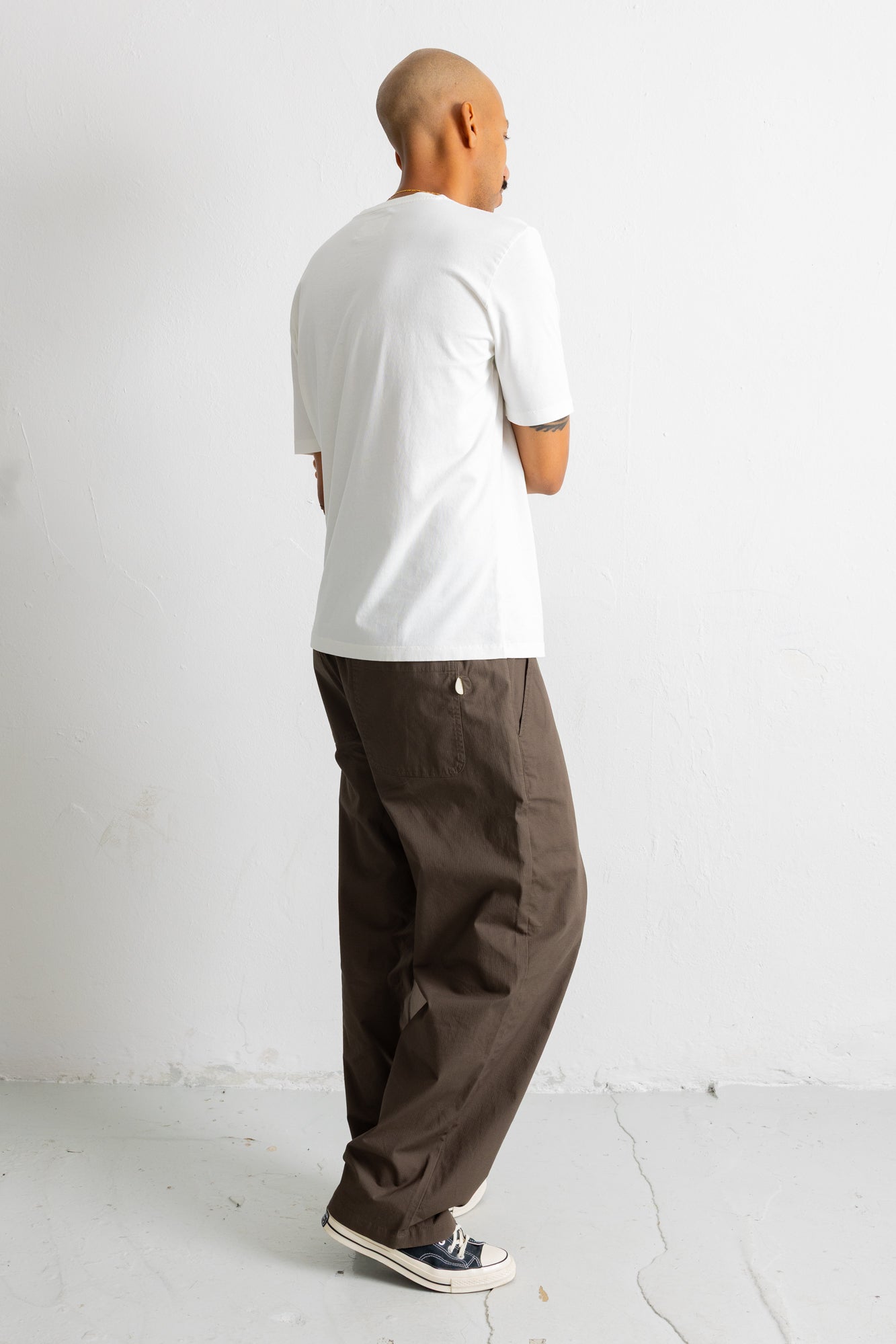 A man shown from the back wearing a white t-shirt, loose brown pants, and dark sneakers, standing against a plain white wall.