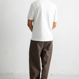 A man viewed from behind wearing a plain white short-sleeve t-shirt and dark brown relaxed-fit pants, standing against a white studio wall.