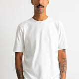 A man with a mustache and tattoos wearing a plain white short-sleeve crewneck t-shirt and brown trousers, standing against a light gray studio background.