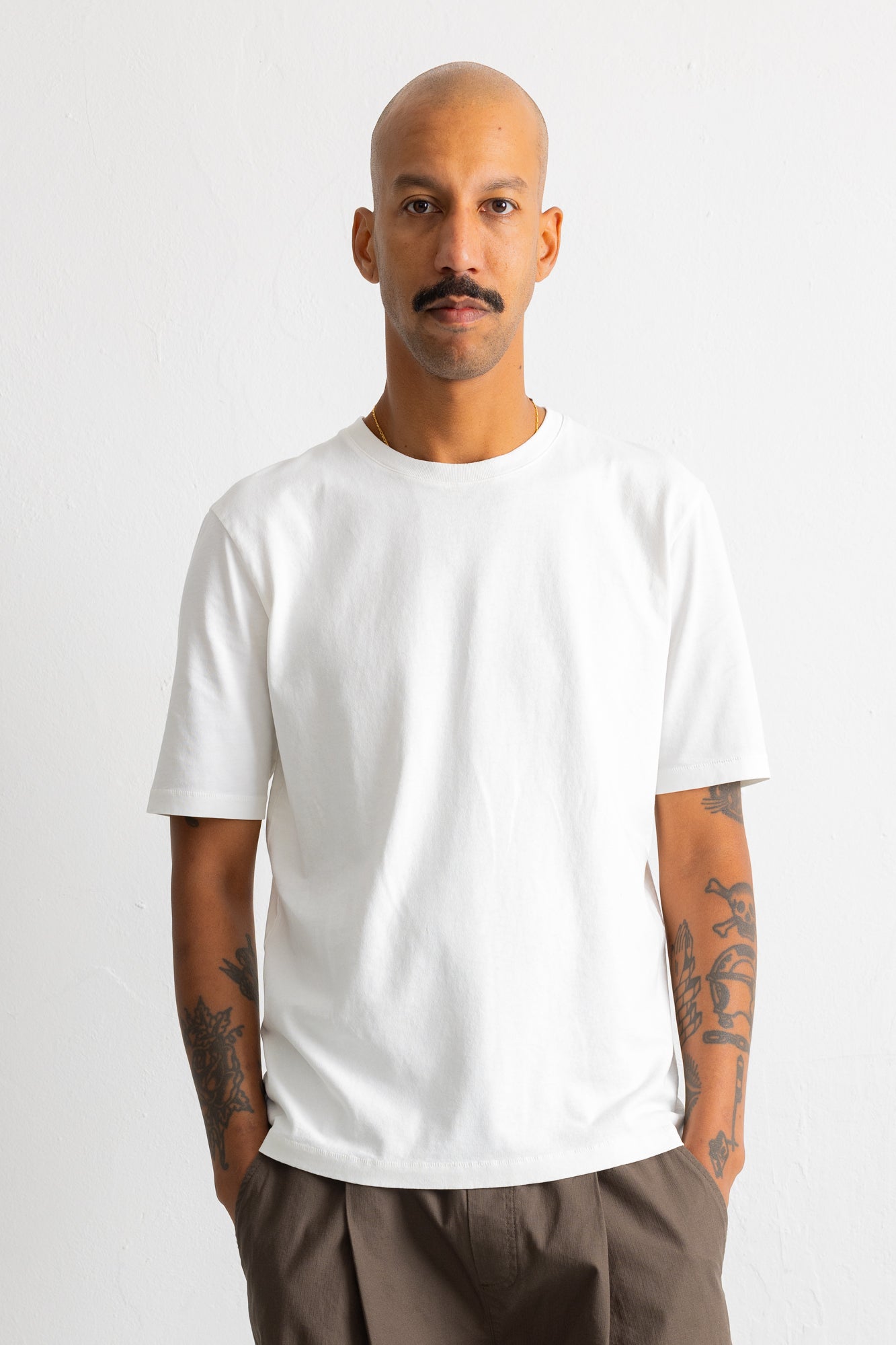 A man with a mustache and tattoos wearing a plain white short-sleeve crewneck t-shirt and brown trousers, standing against a light gray studio background.