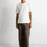 A man wearing a plain white short-sleeve t-shirt, loose dark brown pants, and black-and-white sneakers, standing against a plain white wall.