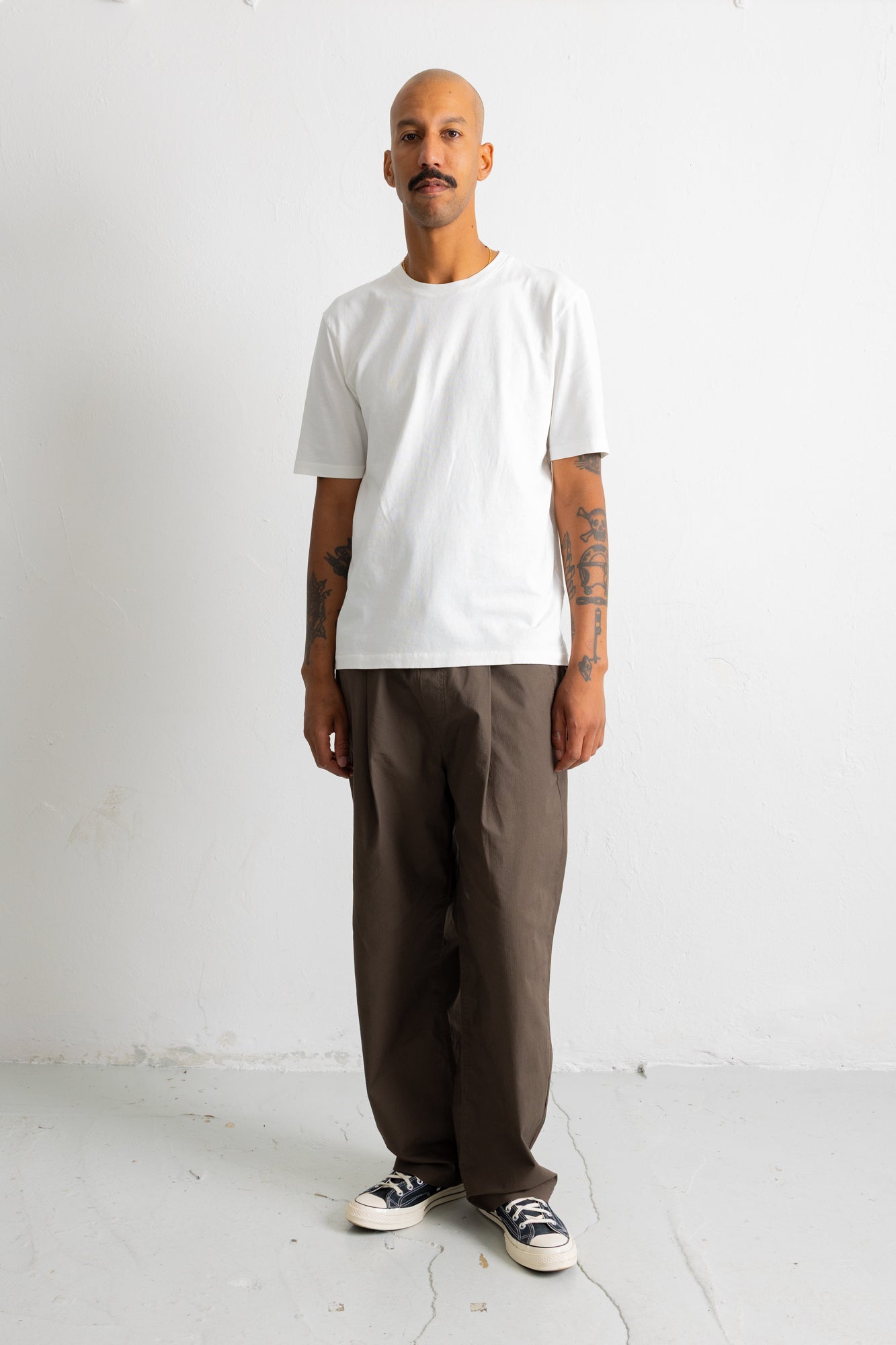 A man wearing a plain white short-sleeve t-shirt, loose dark brown pants, and black-and-white sneakers, standing against a plain white wall.