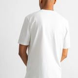 A man viewed from behind wearing a plain white short-sleeve crewneck t-shirt and brown pants, standing against a light gray studio background.