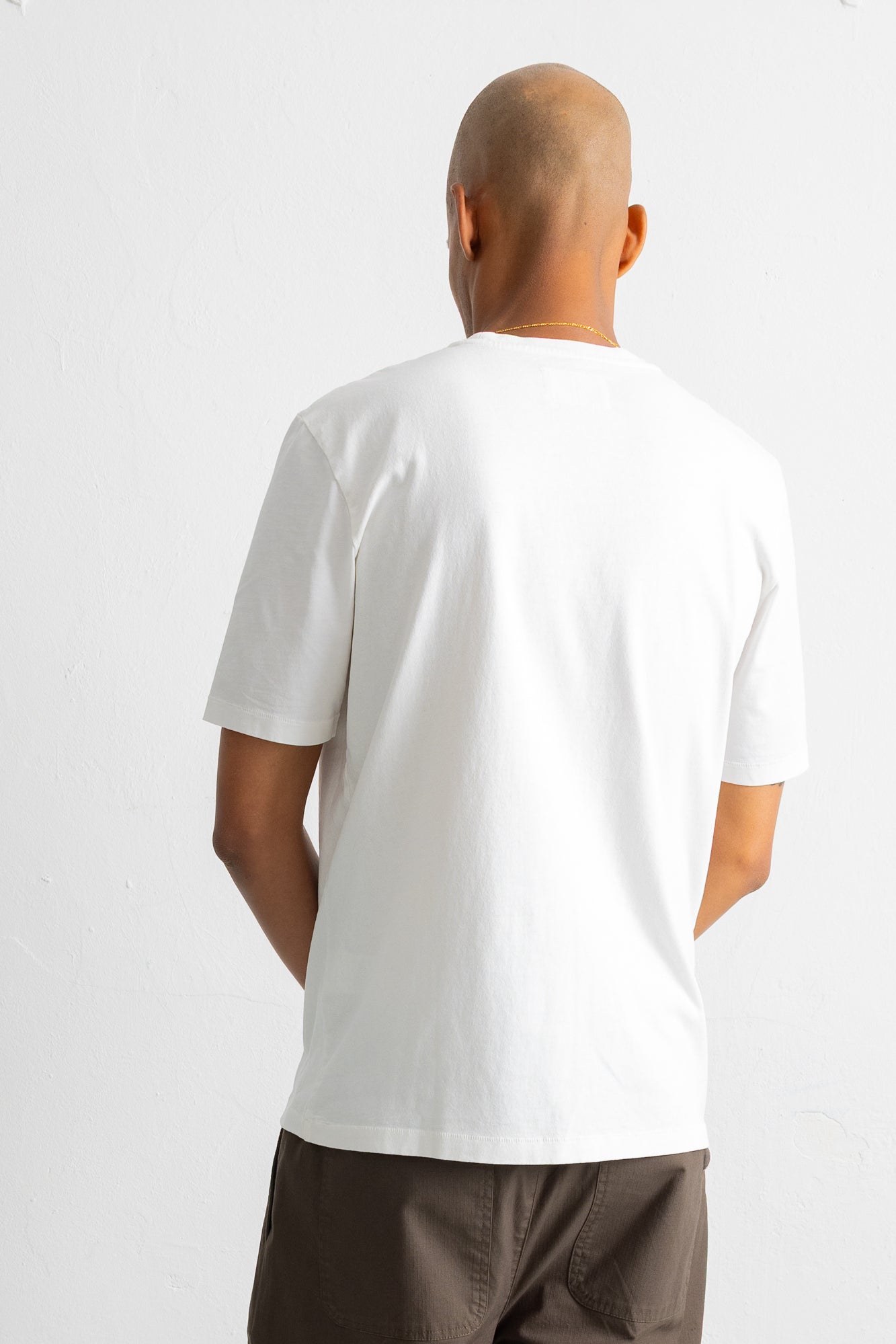 A man viewed from behind wearing a plain white short-sleeve crewneck t-shirt and brown pants, standing against a light gray studio background.