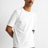 A man wearing a plain white short-sleeve crewneck t-shirt and brown trousers, standing against a light gray studio background.