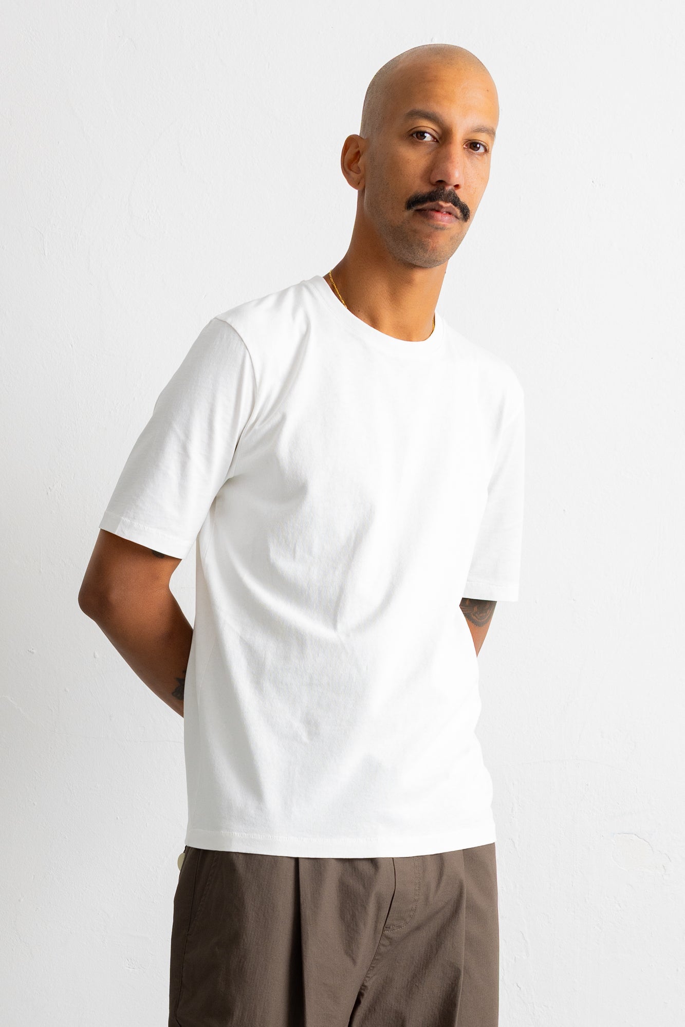 A man wearing a plain white short-sleeve crewneck t-shirt and brown trousers, standing against a light gray studio background.