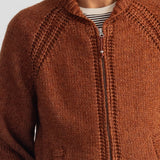 A close-up of a person wearing a rust-orange knit zip-up jacket over a striped shirt, showing the textured knit fabric and collar.