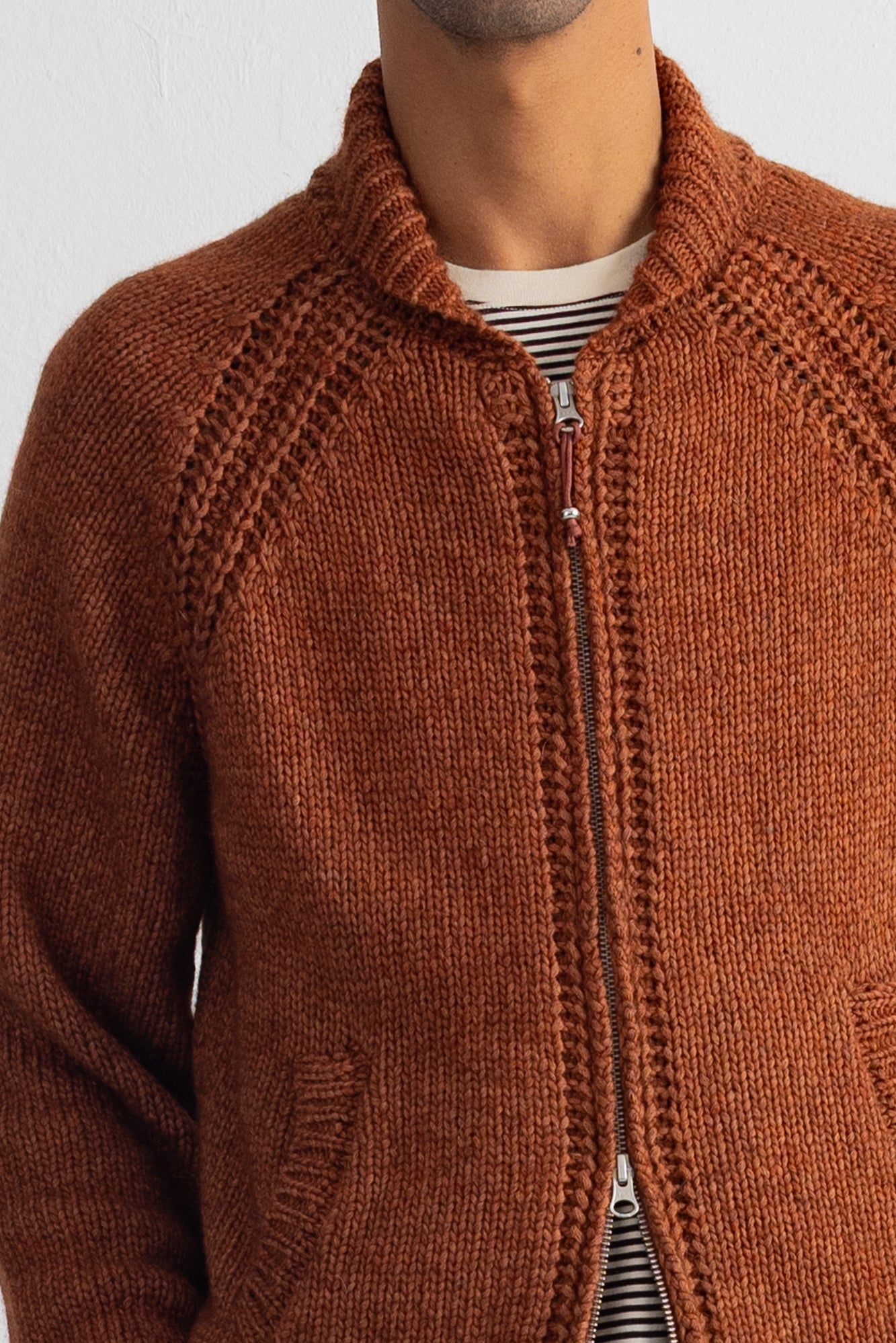 A close-up of a person wearing a rust-orange knit zip-up jacket over a striped shirt, showing the textured knit fabric and collar.