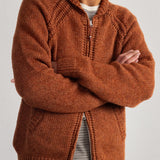 A man wearing a textured rust-orange knit zip-up cardigan over a striped shirt and blue jeans, standing with arms crossed against a plain light gray background.