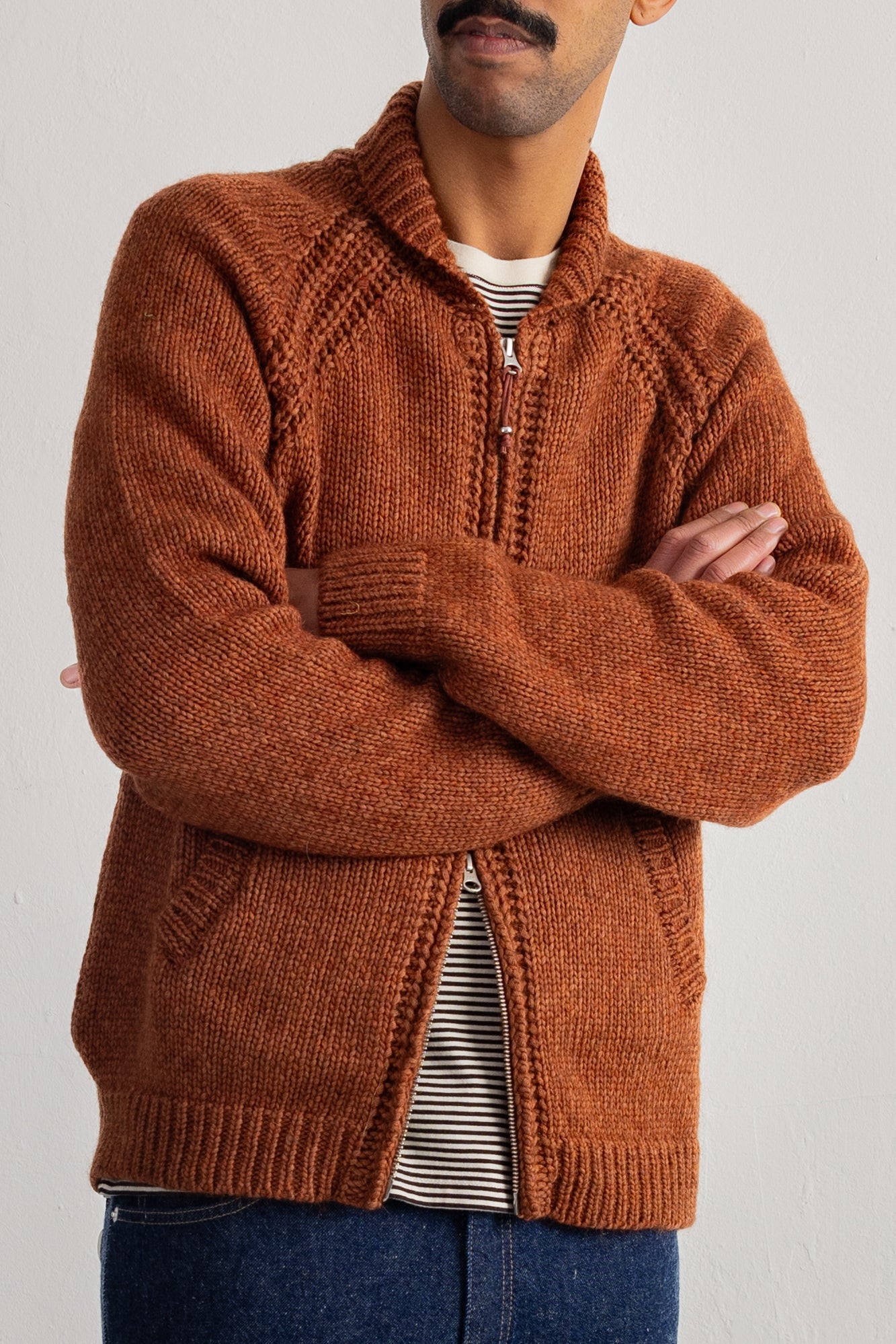 A man wearing a textured rust-orange knit zip-up cardigan over a striped shirt and blue jeans, standing with arms crossed against a plain light gray background.