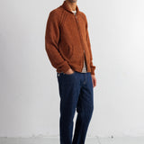 A man wearing a rust-orange knit zip-up cardigan, dark blue jeans, and black shoes, standing against a white studio wall.