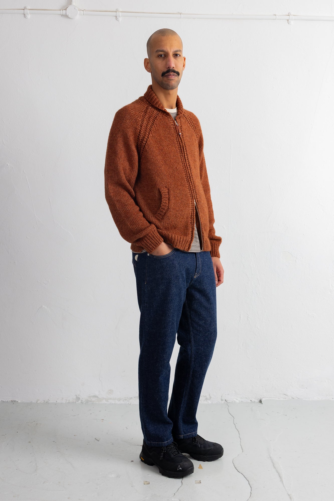 A man wearing a rust-orange knit zip-up cardigan, dark blue jeans, and black shoes, standing against a white studio wall.