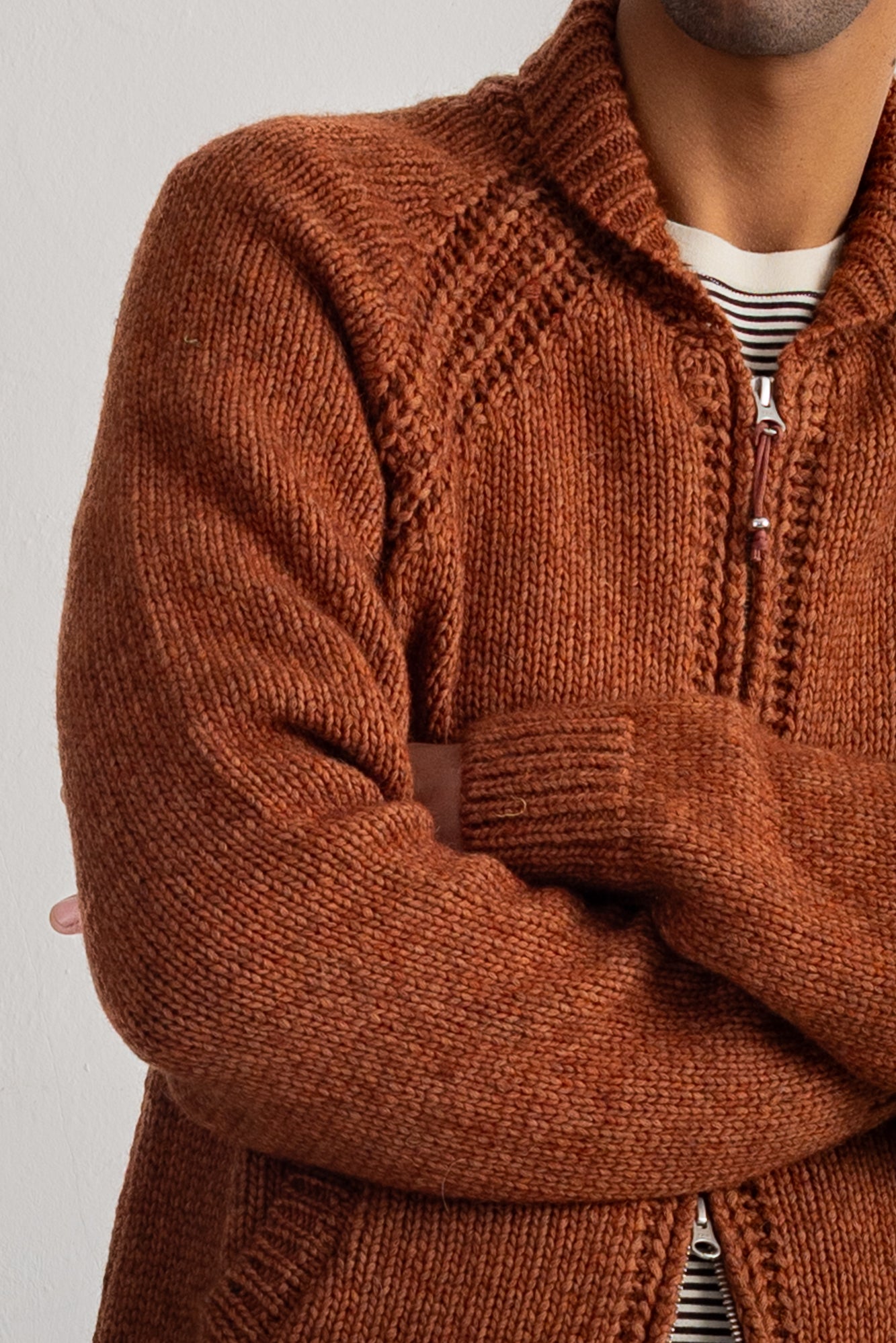A close-up of a man wearing a chunky knit zip-up cardigan in a warm rust brown color, layered over a white and black striped t-shirt.