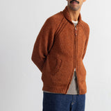 A man wearing a rust-orange knit zip-up cardigan over a striped shirt and blue jeans, standing against a plain white studio background.