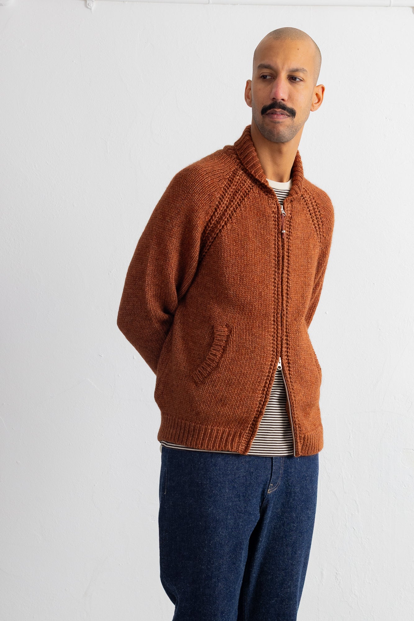 A man wearing a rust-orange knit zip-up cardigan over a striped shirt and blue jeans, standing against a plain white studio background.