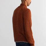 A man shown from the back wearing a rust-brown ribbed knit sweater and dark blue jeans, standing against a plain white wall.