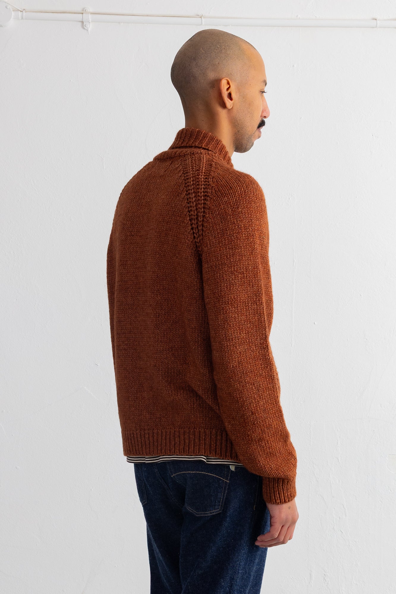 A man shown from the back wearing a rust-brown ribbed knit sweater and dark blue jeans, standing against a plain white wall.