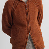 A man wearing a rust-brown knit zip-up cardigan over a striped t-shirt and blue jeans, shown from the chin down against a plain white wall.