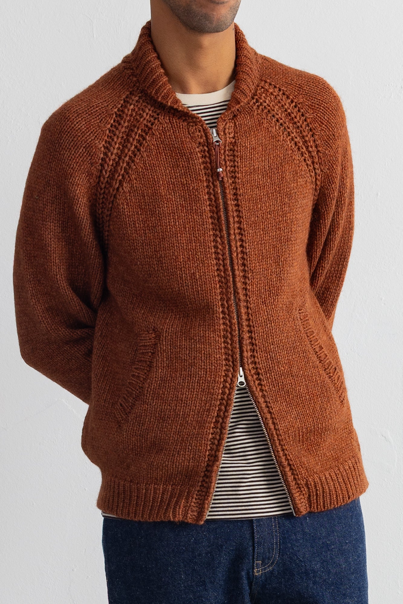 A man wearing a rust-brown knit zip-up cardigan over a striped t-shirt and blue jeans, shown from the chin down against a plain white wall.