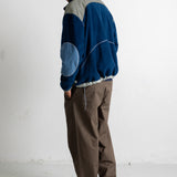 A man wearing a blue and gray color-blocked fleece jacket, brown pants, and black canvas sneakers, standing in profile against a white wall.