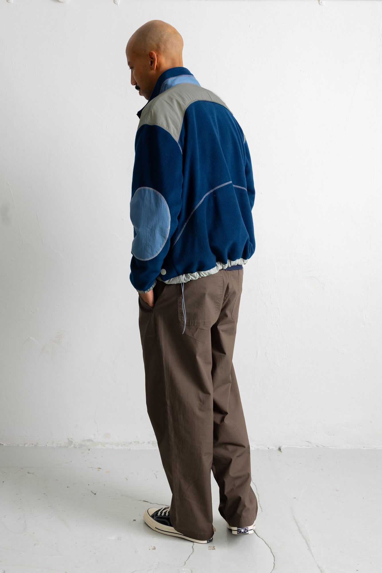 A man wearing a blue and gray color-blocked fleece jacket, brown pants, and black canvas sneakers, standing in profile against a white wall.