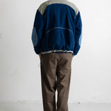 A man viewed from behind wearing a blue and gray color-blocked track jacket and brown pants, standing against a white wall.