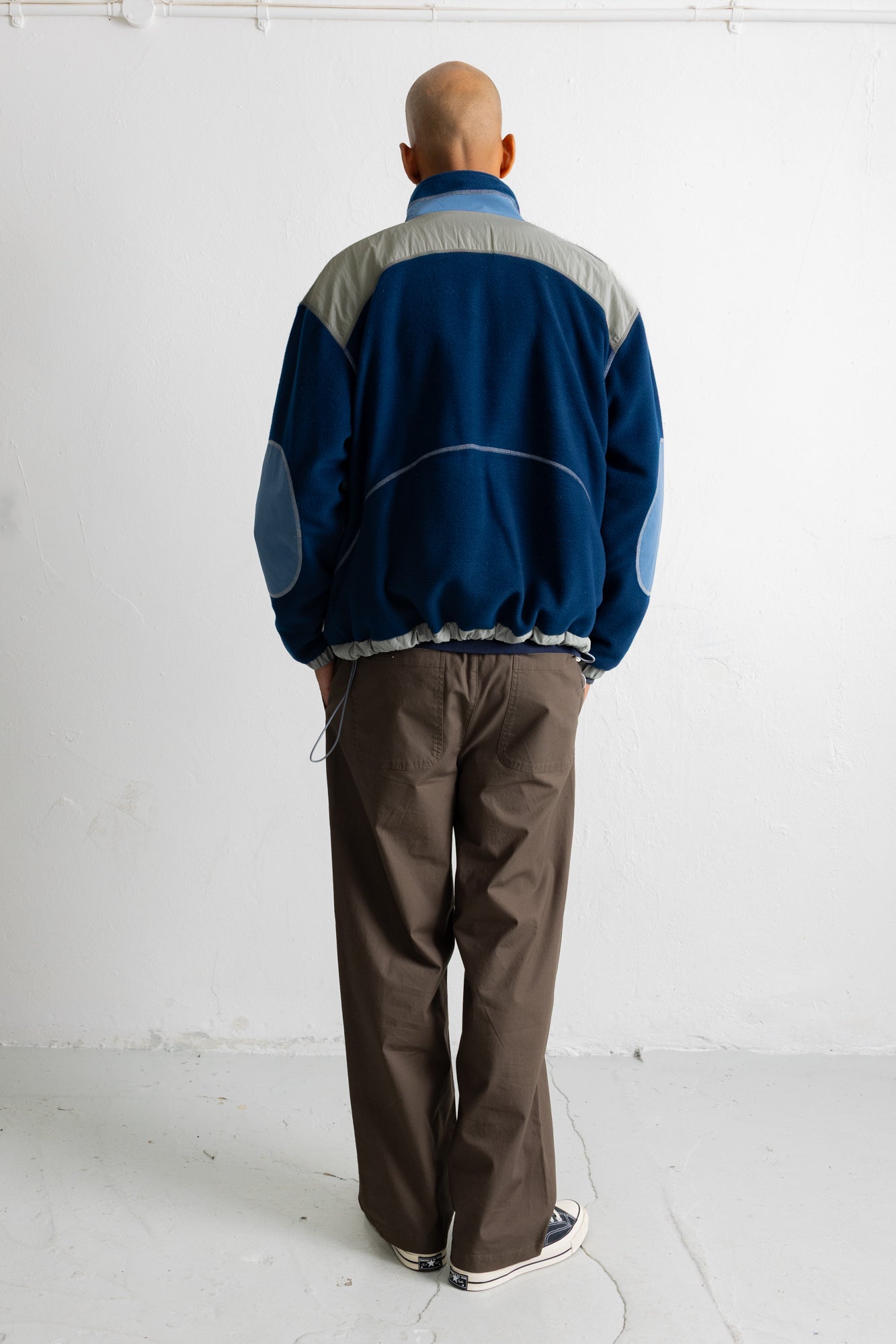 A man viewed from behind wearing a blue and gray color-blocked track jacket and brown pants, standing against a white wall.