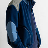 A man wearing a navy blue fleece jacket with light blue and gray color-blocked panels on the shoulders and elbows, paired with brown pants, shown from a side profile against a white wall.