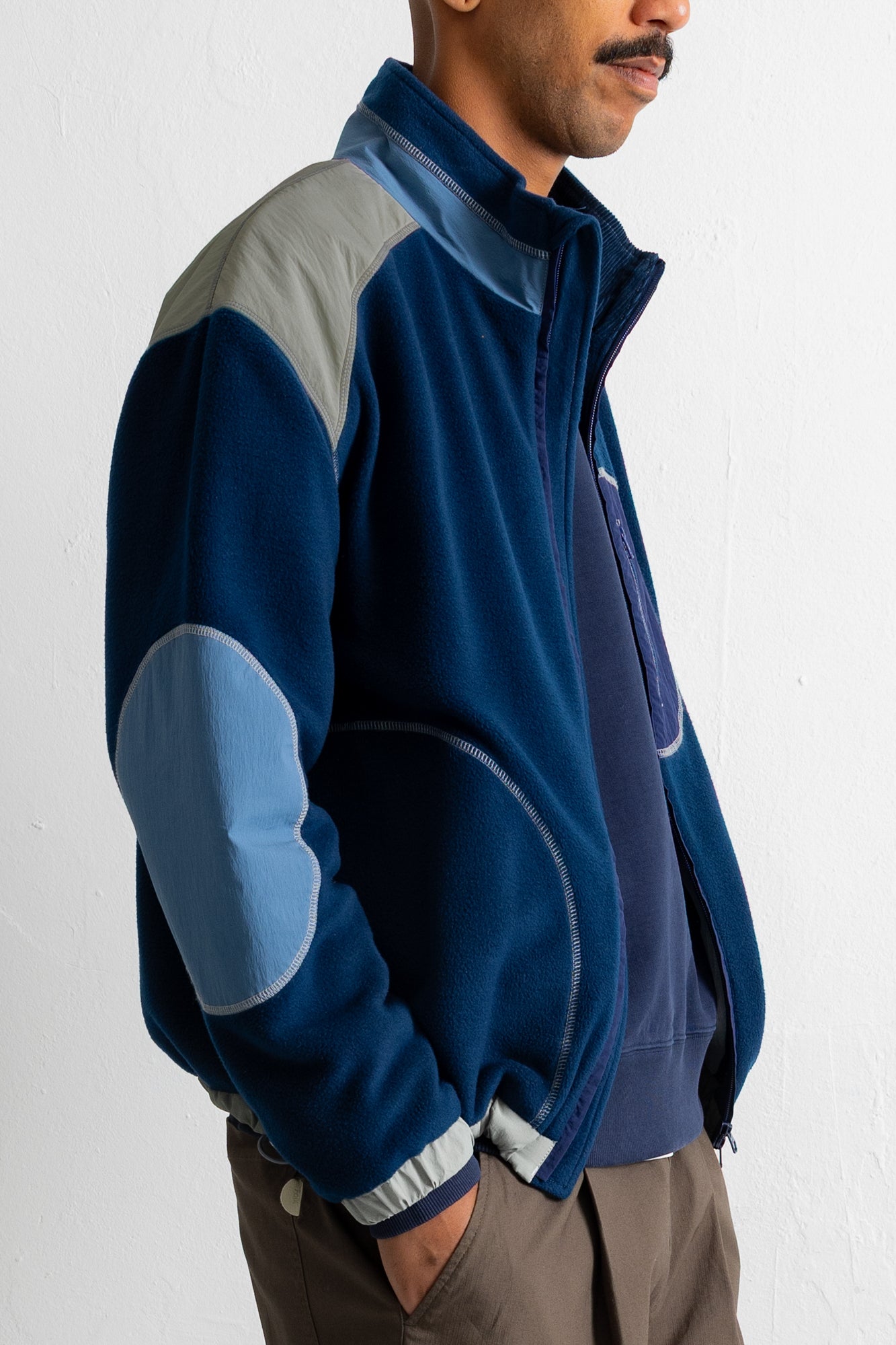A man wearing a navy blue fleece jacket with light blue and gray color-blocked panels on the shoulders and elbows, paired with brown pants, shown from a side profile against a white wall.