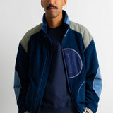 A man wearing a navy blue zip-up jacket with light gray shoulder panels and light blue elbow patches, layered over a dark navy crewneck sweatshirt and brown pants, standing against a plain white wall.