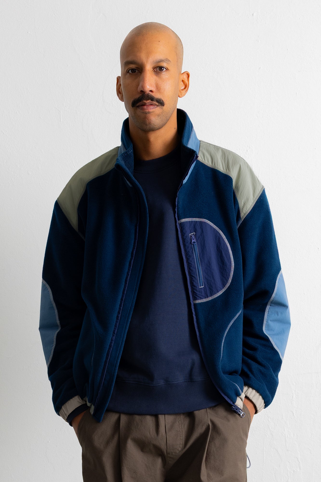 A man wearing a navy blue zip-up jacket with light gray shoulder panels and light blue elbow patches, layered over a dark navy crewneck sweatshirt and brown pants, standing against a plain white wall.