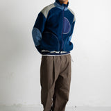 A man wearing a blue and gray patchwork zip-up jacket, brown pants, and black-and-white sneakers, standing against a white wall.