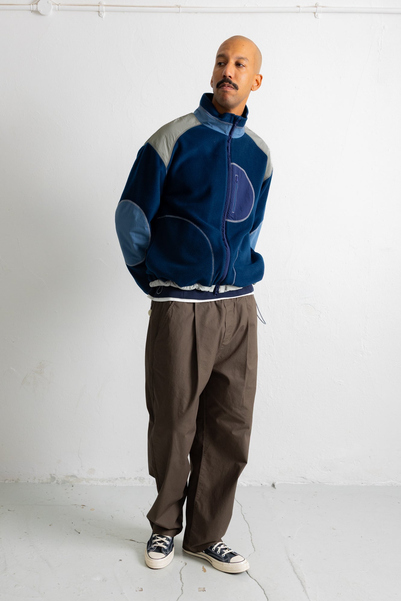 A man wearing a blue and gray patchwork zip-up jacket, brown pants, and black-and-white sneakers, standing against a white wall.