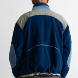 A man viewed from behind wearing a blue and gray color-blocked fleece jacket with brown pants, standing against a plain white wall.