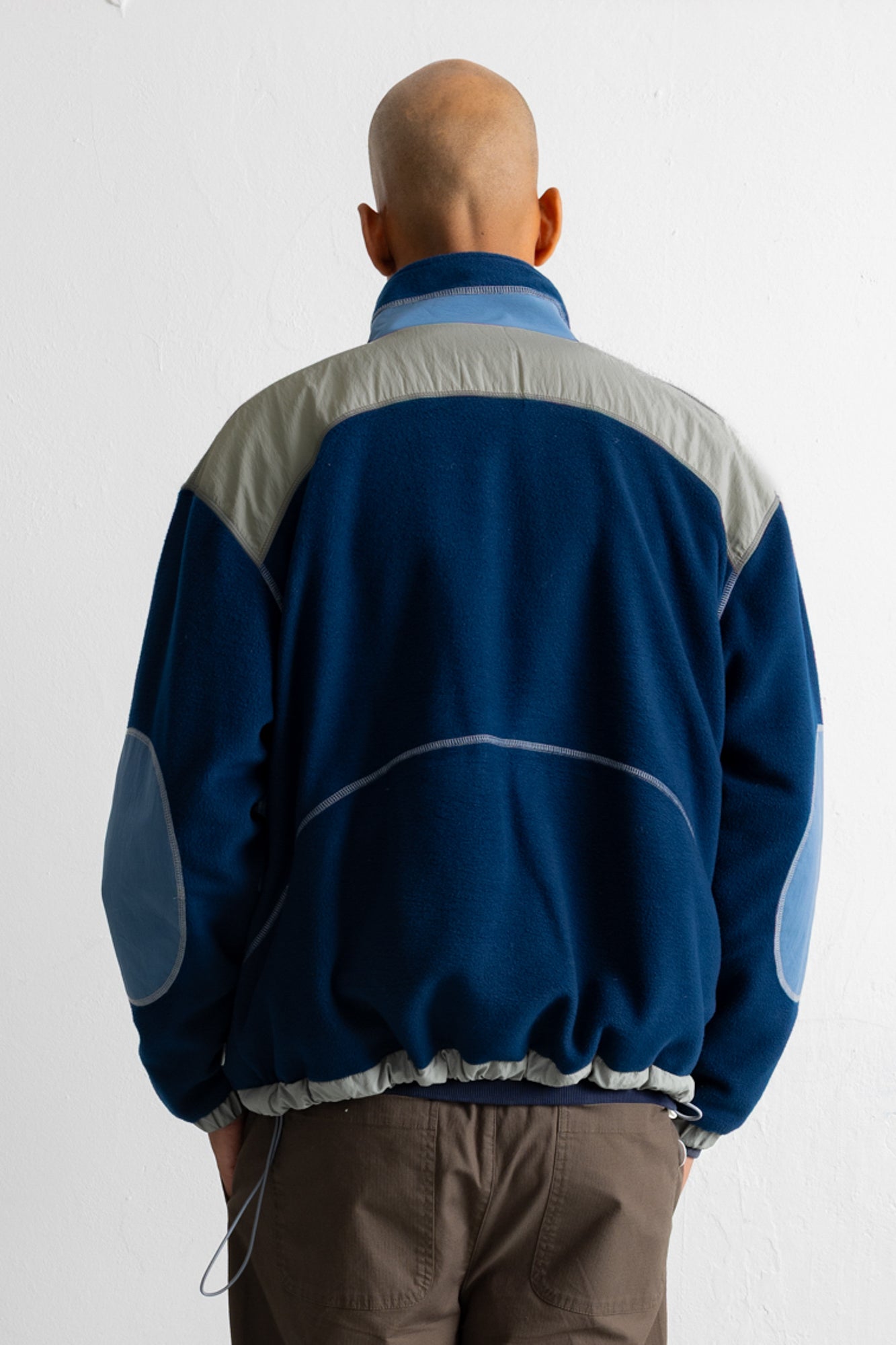A man viewed from behind wearing a blue and gray color-blocked fleece jacket with brown pants, standing against a plain white wall.