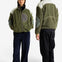 A man and a woman modeling an olive green fleece jacket with contrasting white shoulder panels, gray elbow patches, and a gray zippered chest pocket, shown from both front and back views against a plain white studio background.
