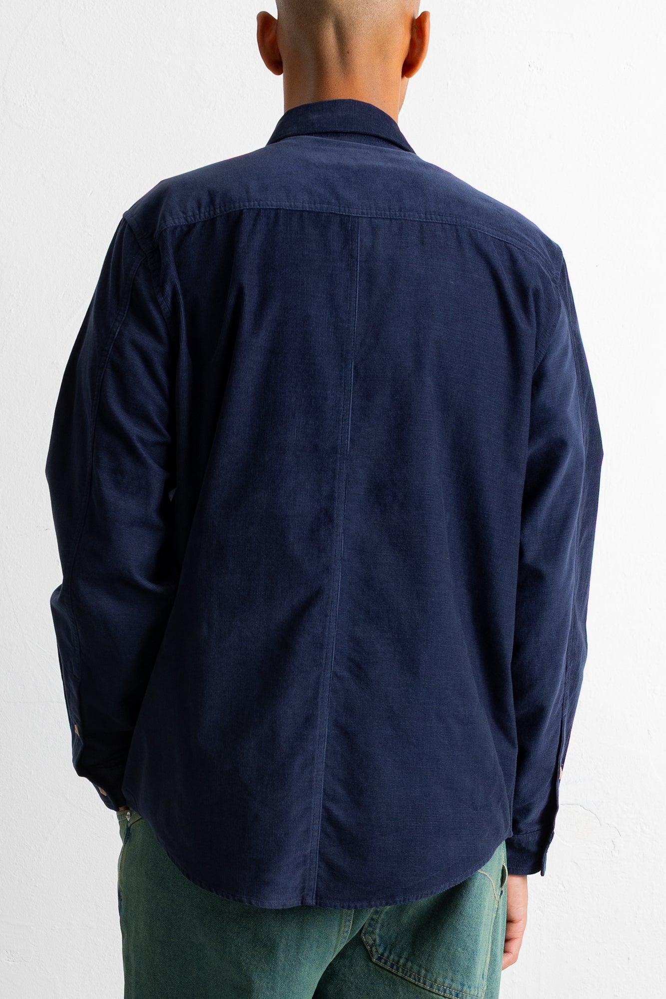 A man viewed from behind wearing a dark navy blue long-sleeve button-down shirt and green pants, against a plain white background.