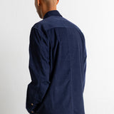 A man viewed from the back wearing a dark navy blue corduroy shirt and green pants, standing against a plain white wall.