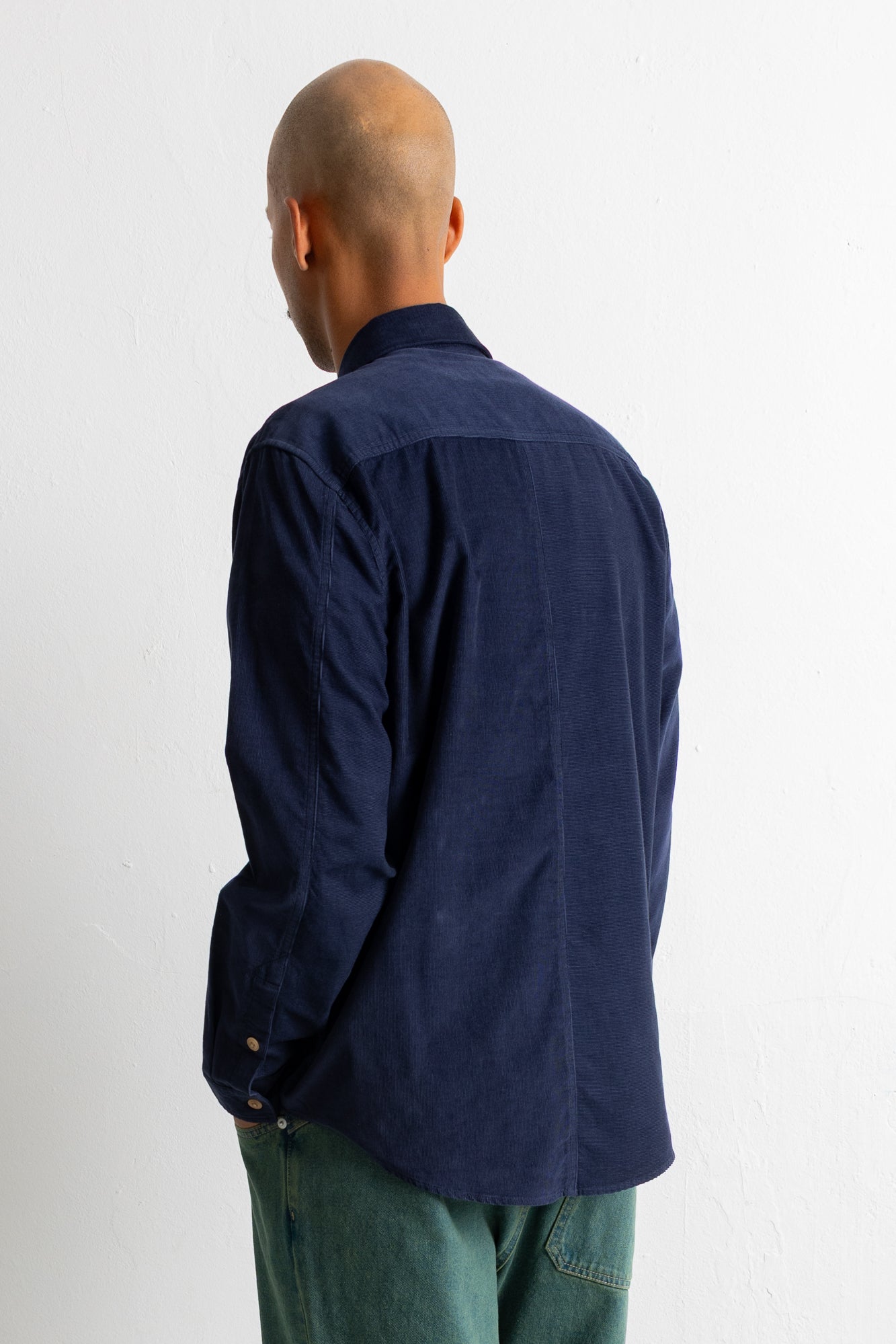 A man viewed from the back wearing a dark navy blue corduroy shirt and green pants, standing against a plain white wall.