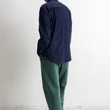 A man standing with his back to the camera, wearing a dark navy blue long-sleeve button-up shirt, green trousers, and black canvas sneakers against a plain white wall.