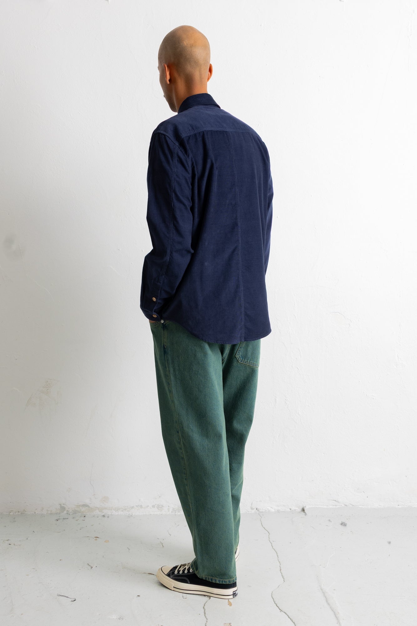 A man standing with his back to the camera, wearing a dark navy blue long-sleeve button-up shirt, green trousers, and black canvas sneakers against a plain white wall.