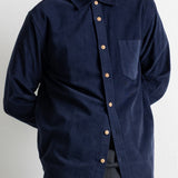 A man wearing a dark navy blue corduroy button-down shirt with a chest pocket and light-colored buttons, paired with green jeans.
