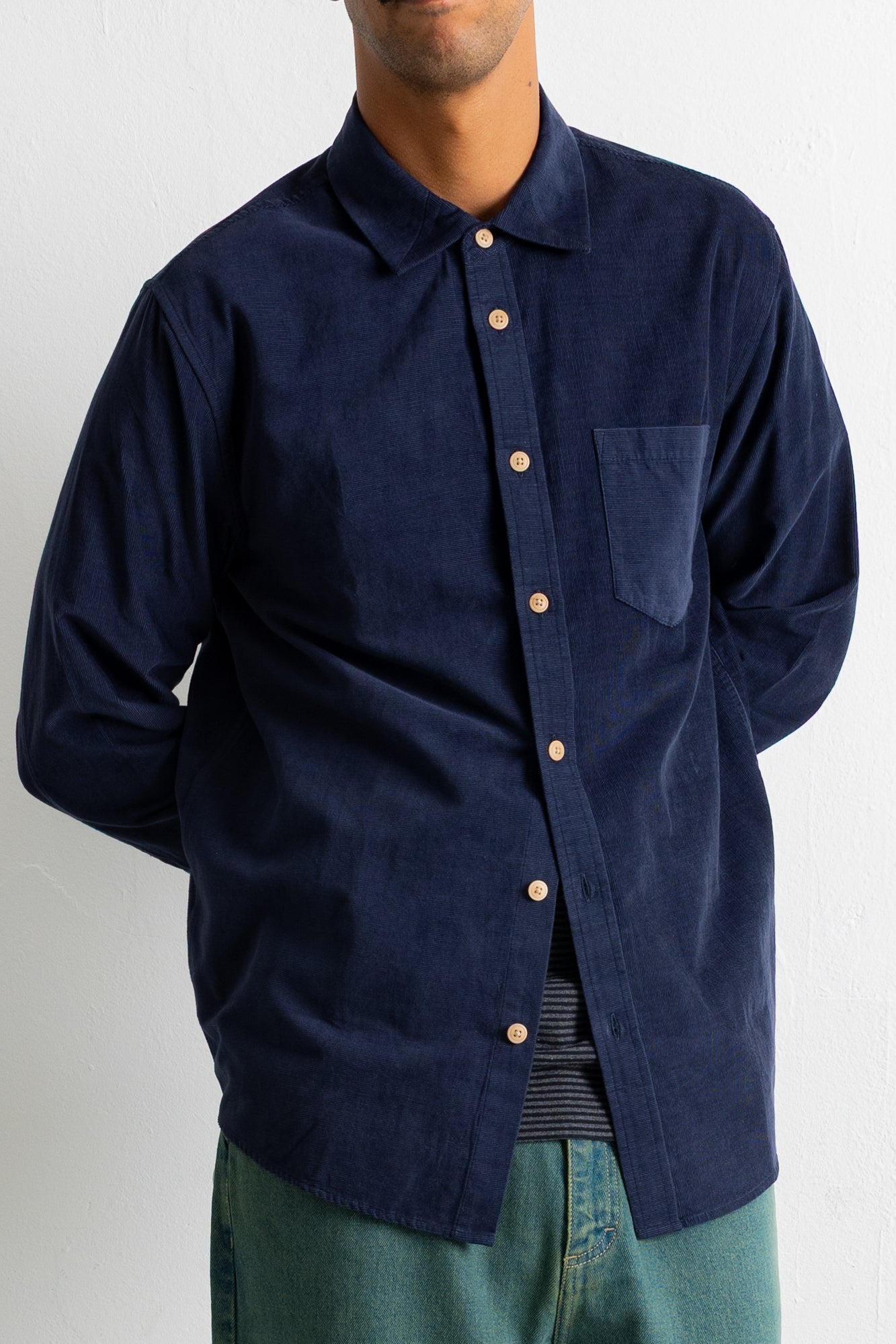 A man wearing a dark navy blue corduroy button-down shirt with a chest pocket and light-colored buttons, paired with green jeans.