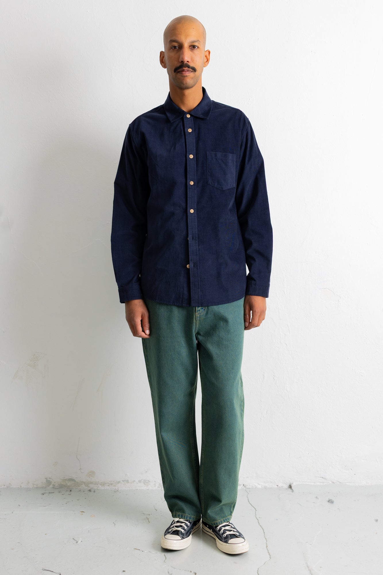 A man wearing a dark navy blue long-sleeve button-up shirt, green pants, and black-and-white sneakers, standing against a plain white wall.