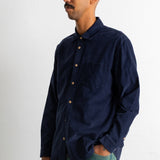 A man wearing a dark navy blue long-sleeve button-down shirt and green pants, standing against a plain white wall.