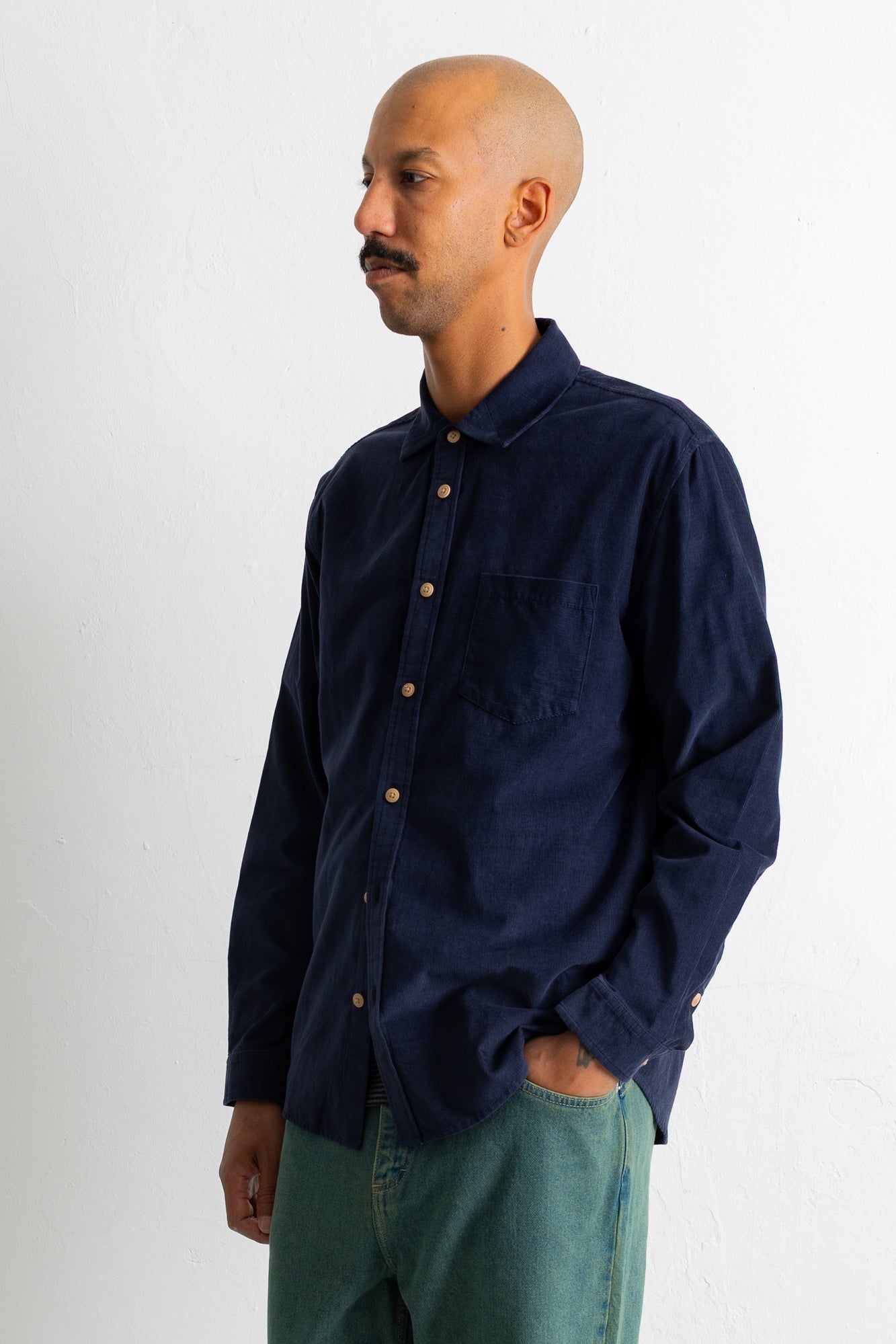 A man wearing a dark navy blue long-sleeve button-down shirt and green pants, standing against a plain white wall.