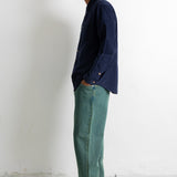 A man stands in profile wearing a dark navy long-sleeve button-up shirt, teal-green trousers, and black canvas sneakers against a plain white wall.